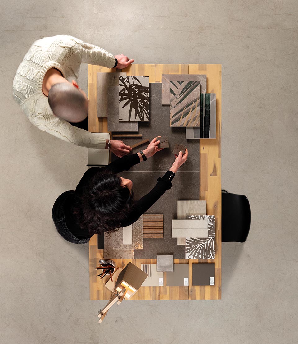 Designer on work table with material moodboard: stone effect stoneware samples, botanical decor in neutral tones, bronze, green and sand.
