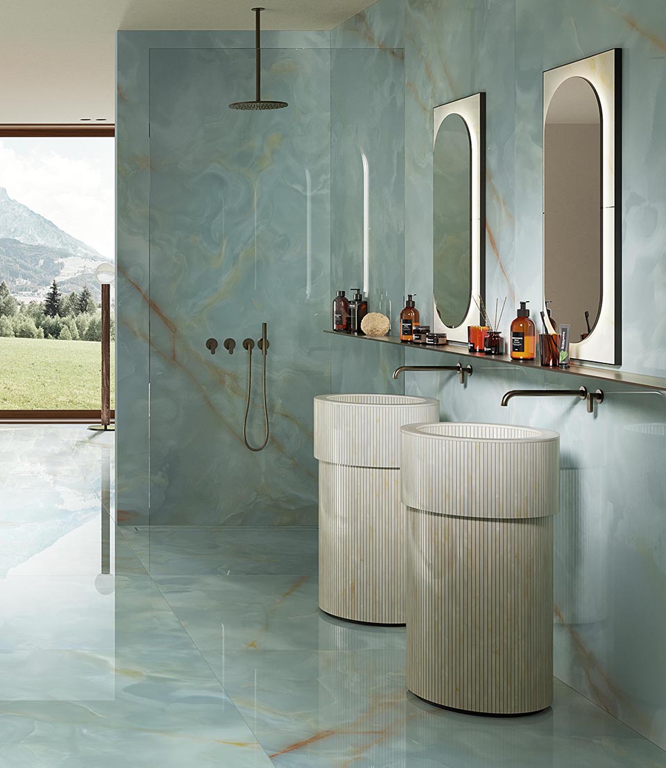 Bathroom with light blue onyx-effect stoneware wall tiles with orange veins, fluted washbasins, bronze fittings, oval mirrors.
