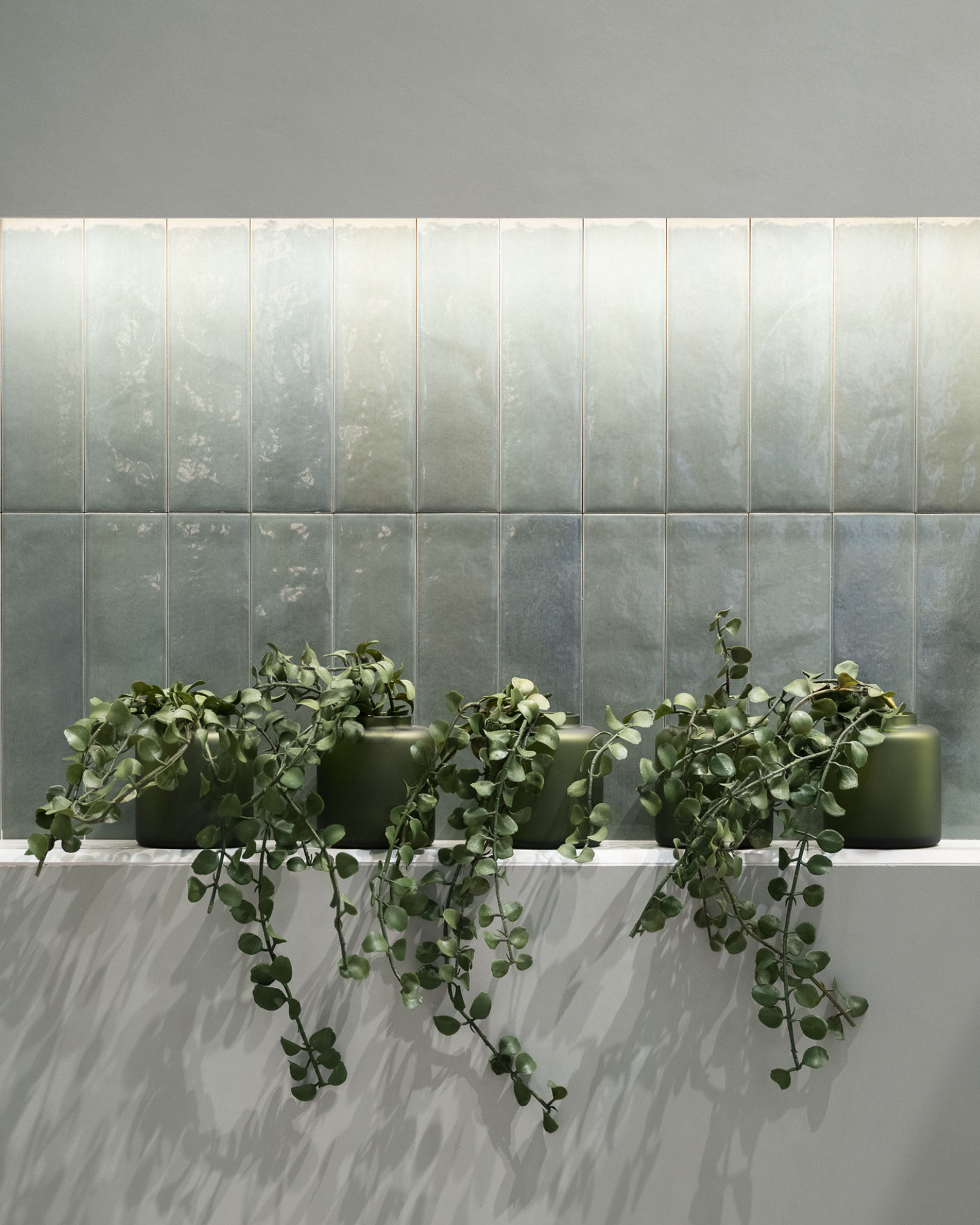 Wall covered with glossy sea green ceramic tiles, decorative plants in green pots on a white shelf with a minimalist design.