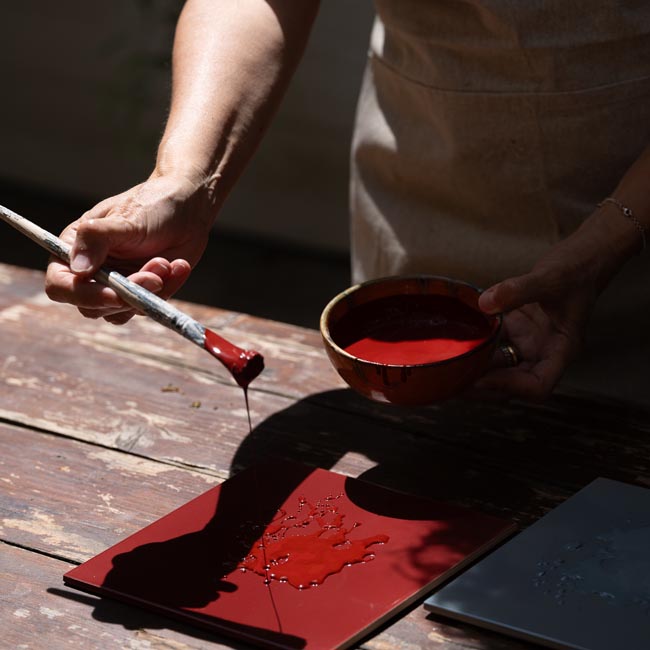 Craftsman applying glossy red paint to ceramic tile with brush and terracotta bowl.