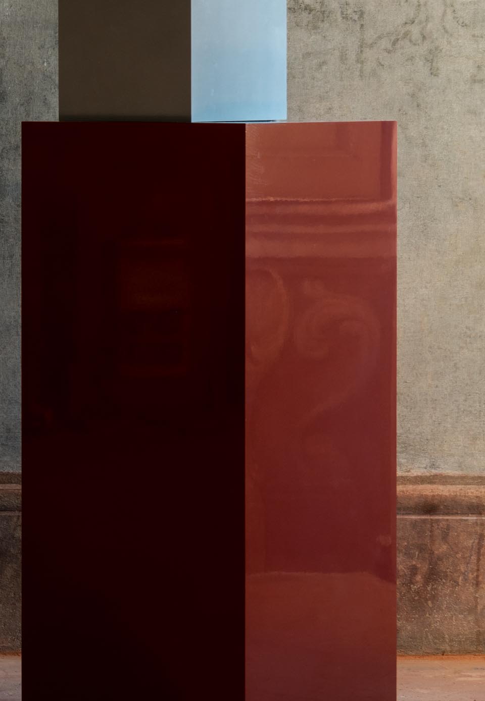 Ceramic volumes with a glossy bright red and matte blue finish, on a neutral textured background with decorative details.