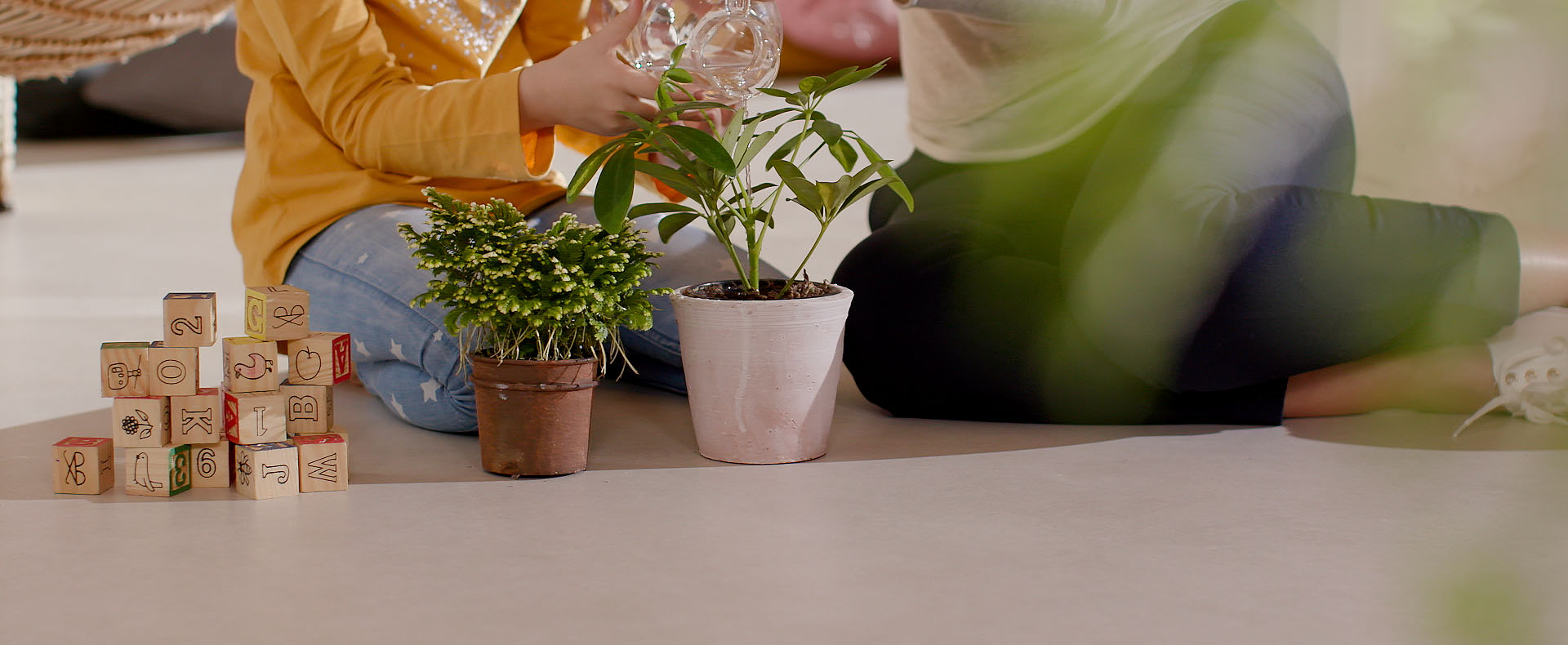 Two children sitting on beige stoneware floor, tending potted plants and playing with wooden cubes.