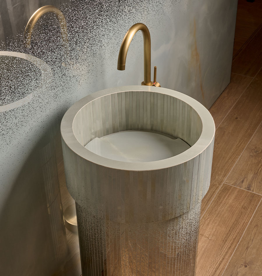 Light ceramic cylindrical washbasin with vertical strip cladding, gold brass tap and wood-effect flooring.
