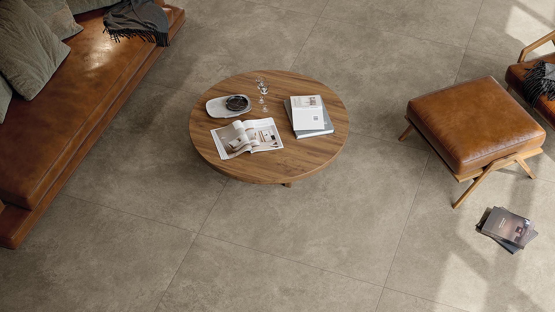 Living room with grey stoneware effect floor, leather sofa and armchair, round central table in natural wood.