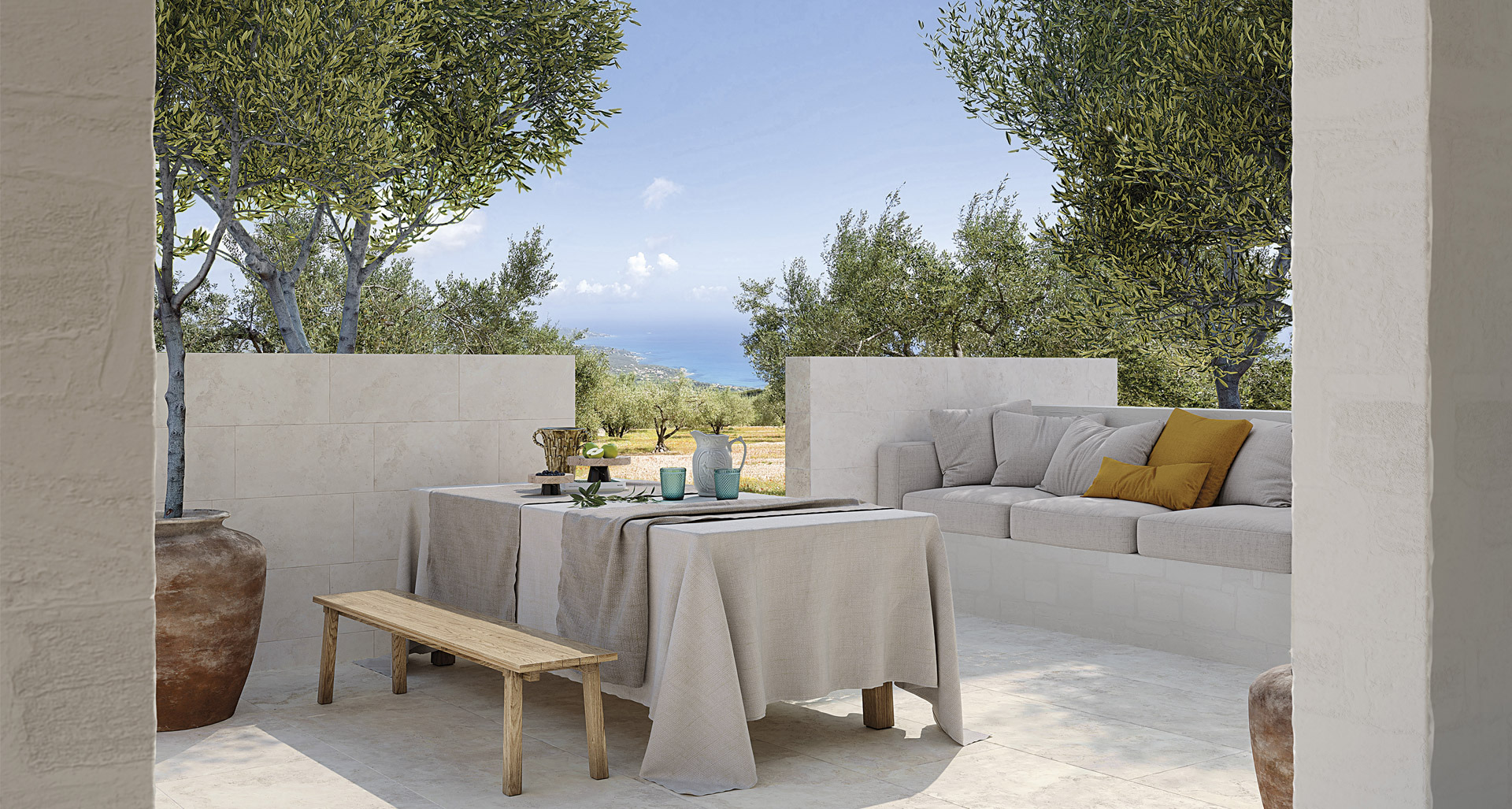 Outdoors with light stoneware effect floor, wood and linen furniture, masonry sofa with cushions and a view of olive trees and the sea.