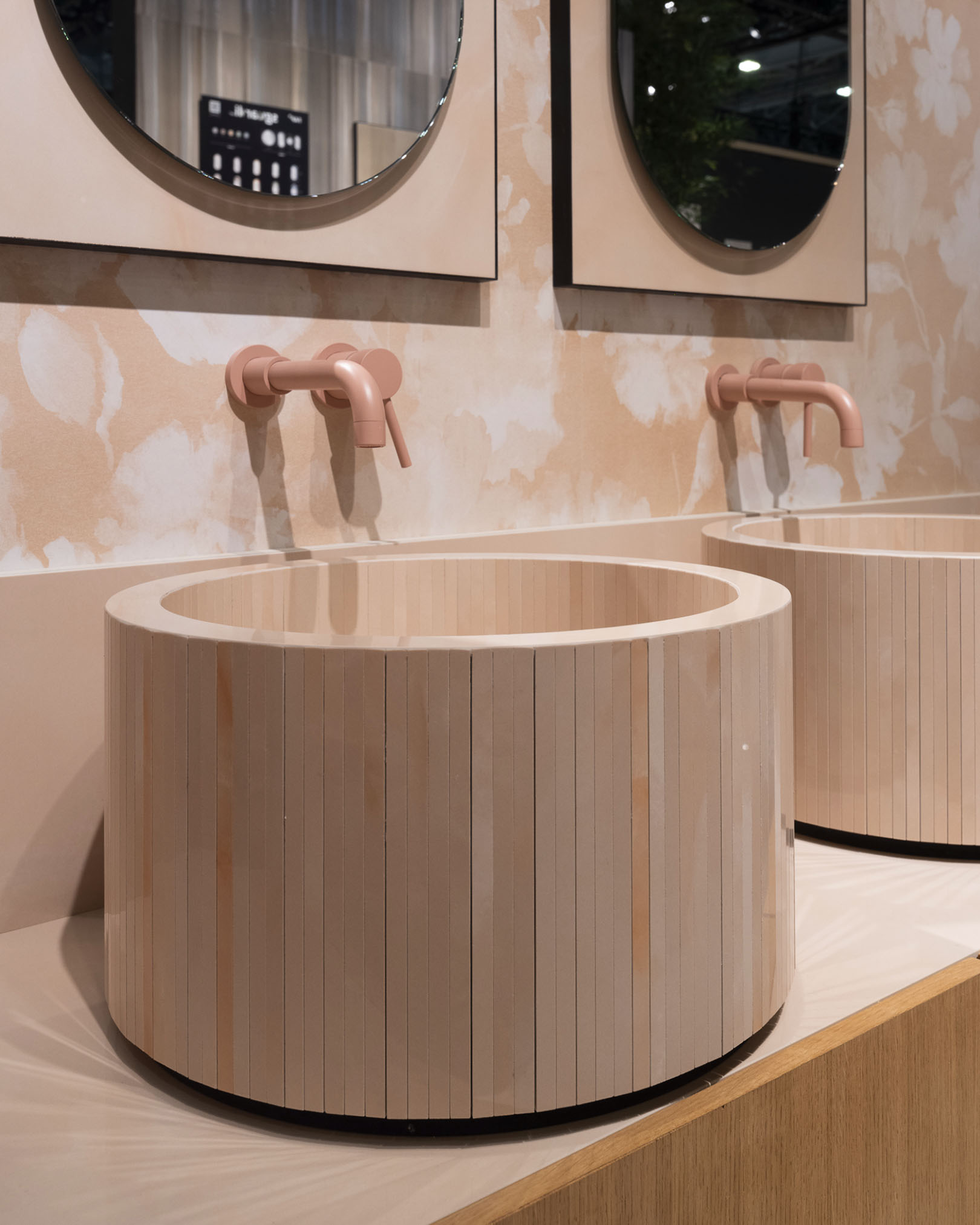 Bathroom with cylindrical washbasin in light wood with vertical slats, matt pink taps and decorative beige floral wall.