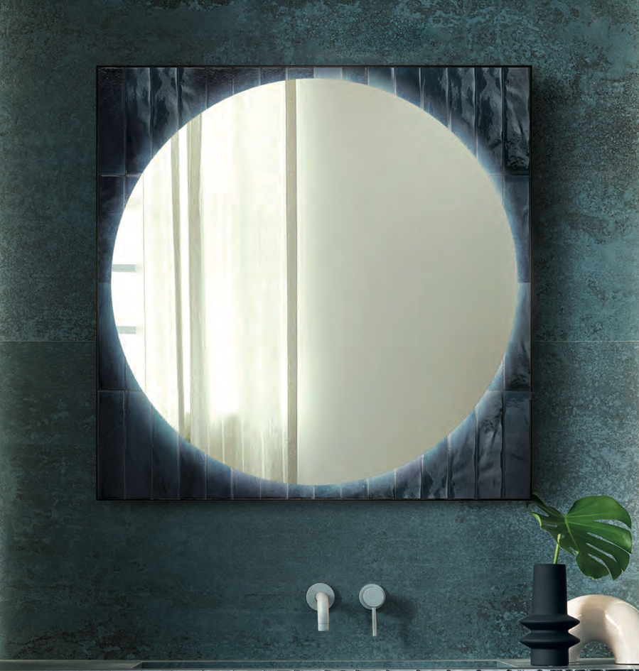 Round mirror framed by glossy blue tiles on a powder blue textured wall, with white washbasin and mixer tap.