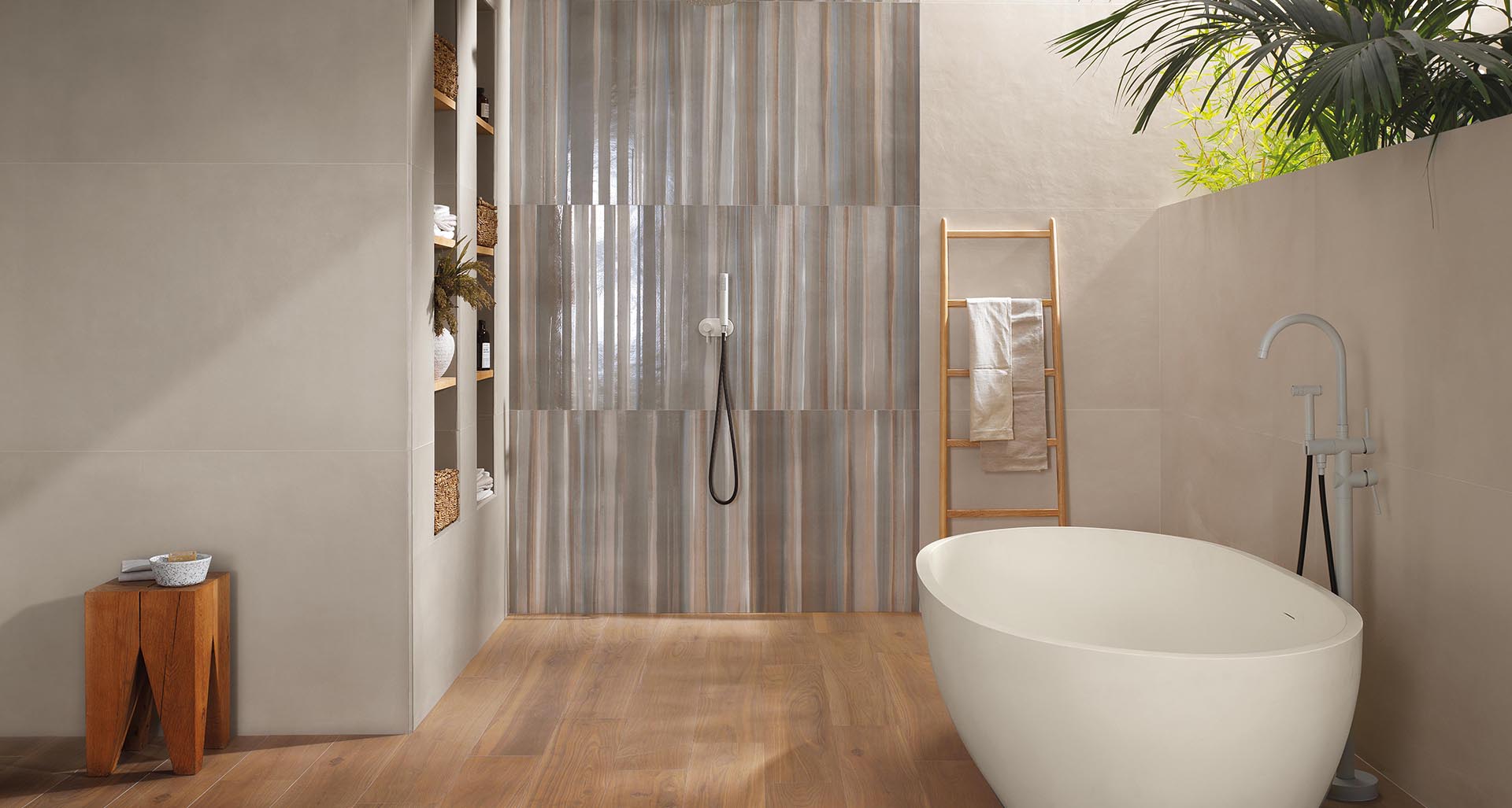 Bathroom with white freestanding bathtub, neutral walls and shower decorated with glossy tiles with vertical stripes in shades of beige and grey.