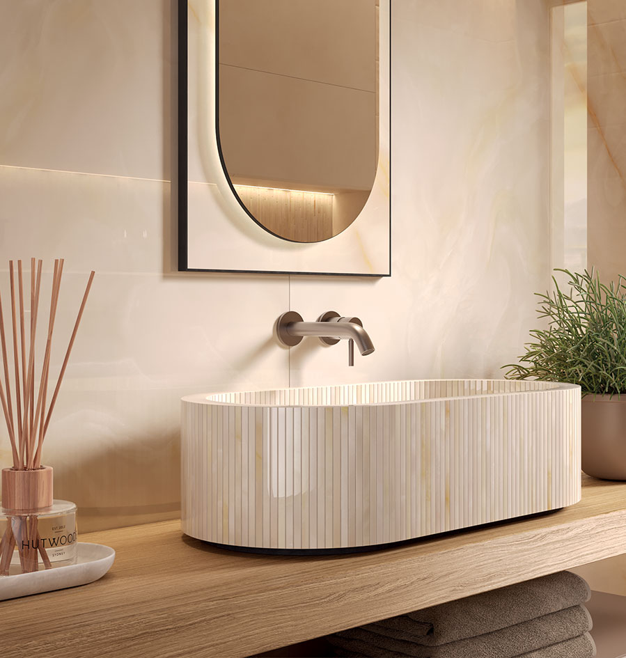 Oval washbasin in glossy ceramic with vertical slatted texture, on natural wood top, backlit mirror and steel tap.
