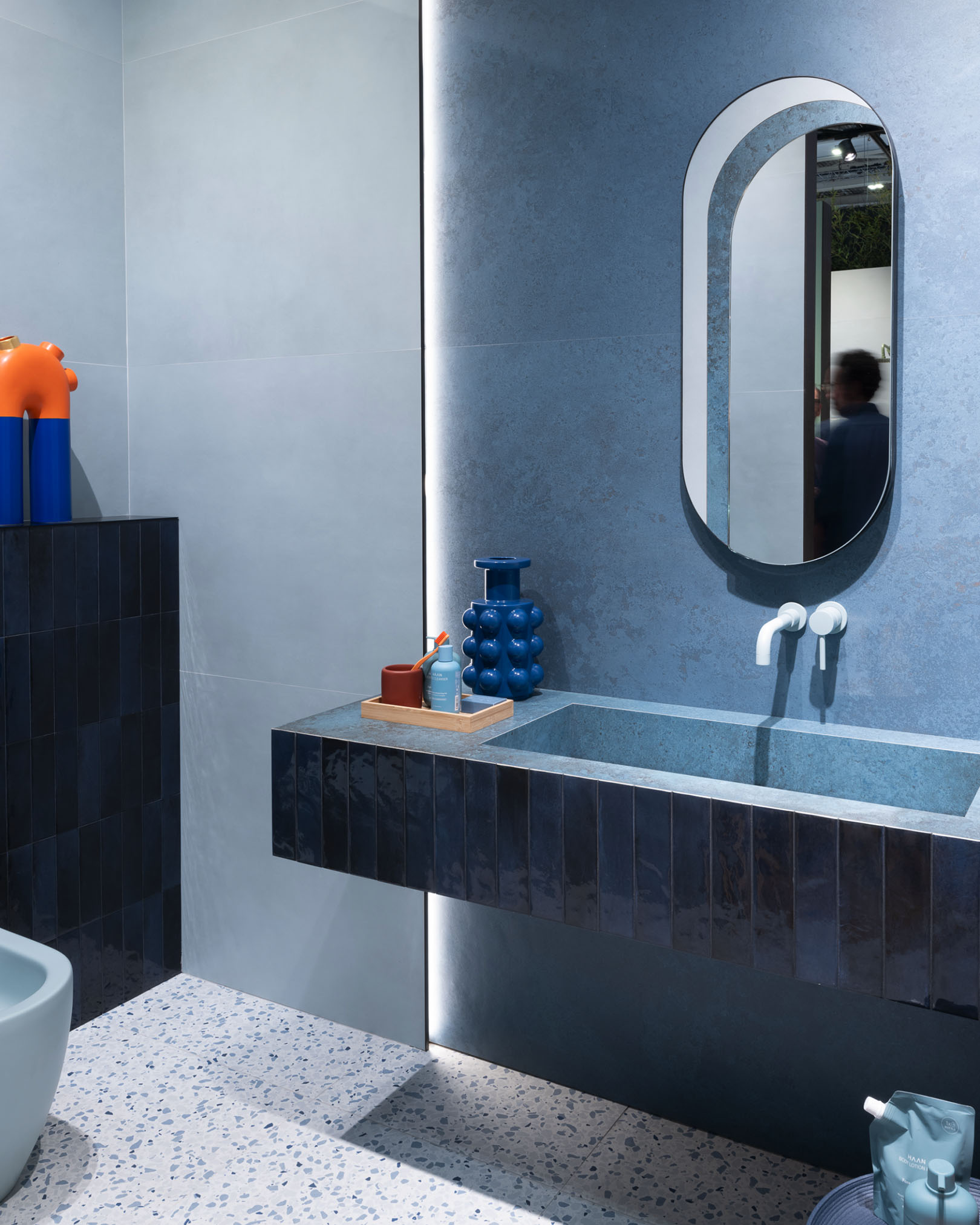 Modern bathroom with blue and grey tiles, wall-mounted glazed stoneware washbasin, terrazzo flooring and backlit oval mirror.