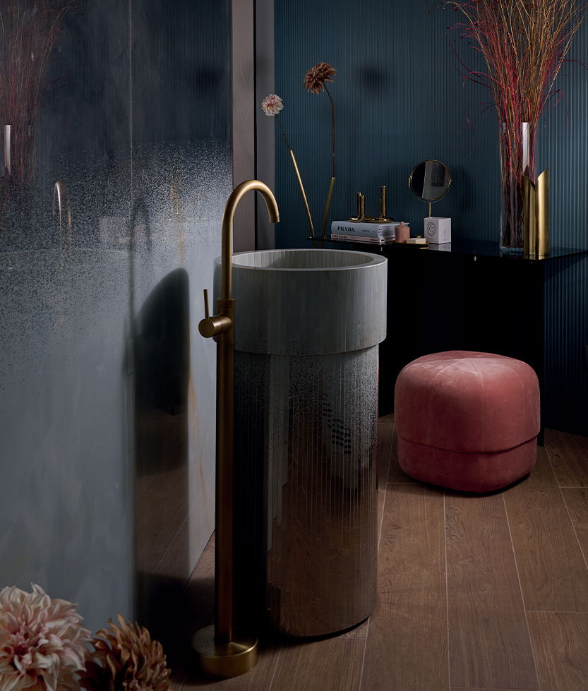 Cylindrical washbasin decorated with brass tap, pink pouf and wood-effect porcelain stoneware walls with a dark texture.