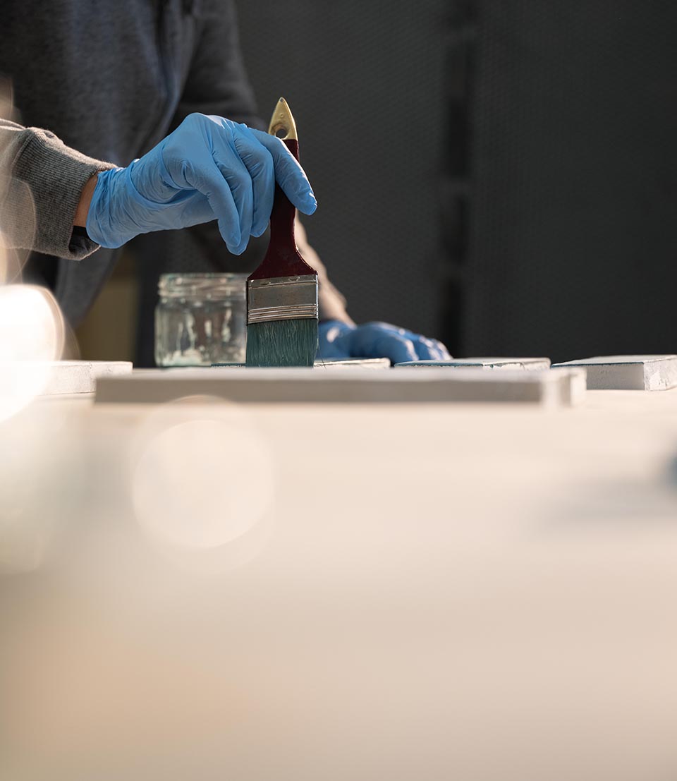 Worker with blue gloves applies glaze on ceramic sample with brush, during color research test in laboratory.
