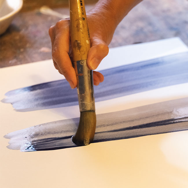 Hand applying blue paint with a flat brush on paper during the creative phase of graphic development.