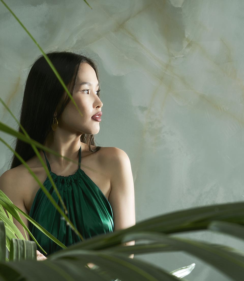 Portrait of an Asian woman on an onyx-effect stoneware background, green hue and golden veining, tropical vegetation in the foreground.