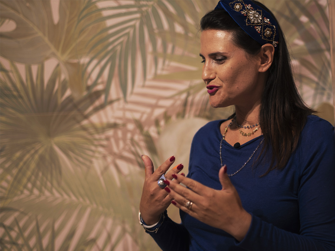 Primo piano di donna in showroom davanti a rivestimento tropicale con foglie verdi, abito blu e dettagli gioiello luminosi.