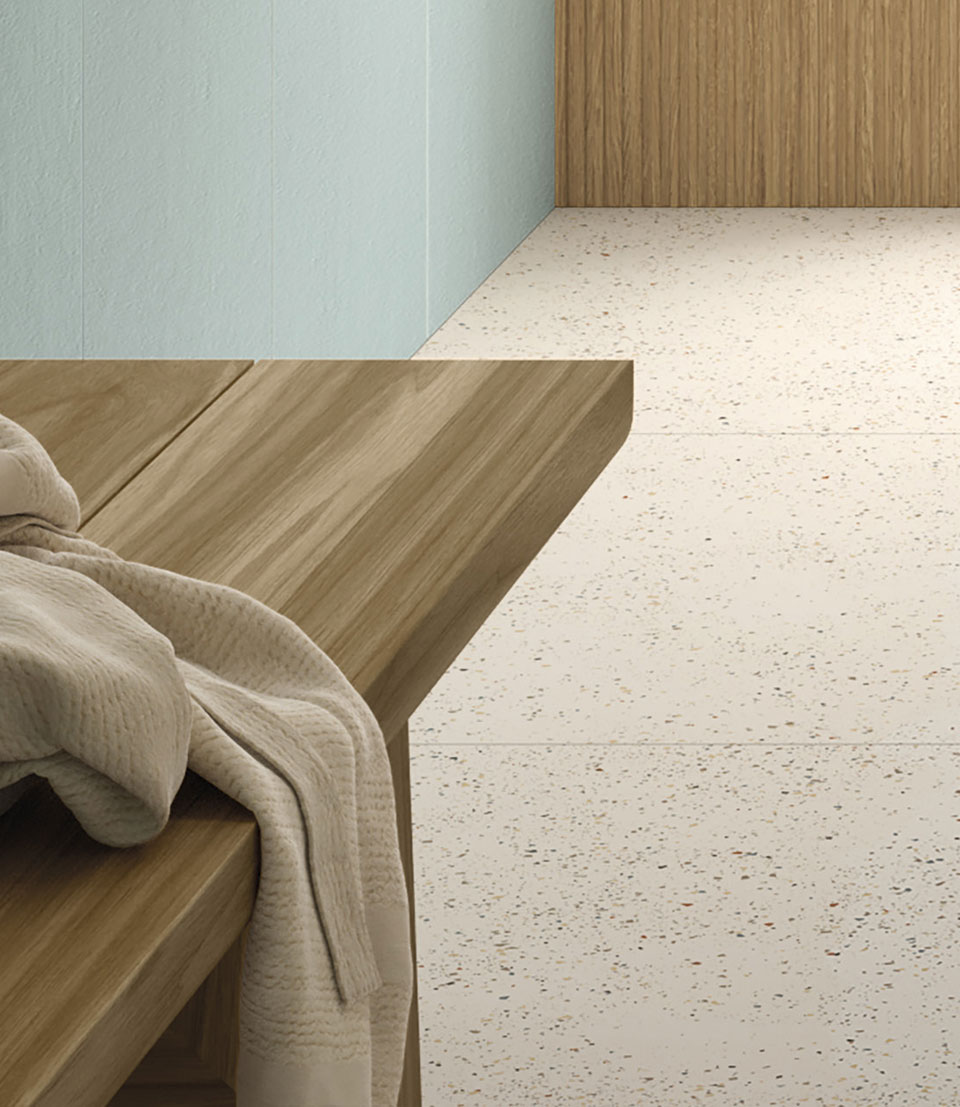 Light terrazzo effect floor, matte blue stoneware wall and natural wood effect stoneware countertop with bathrobe resting on top.
