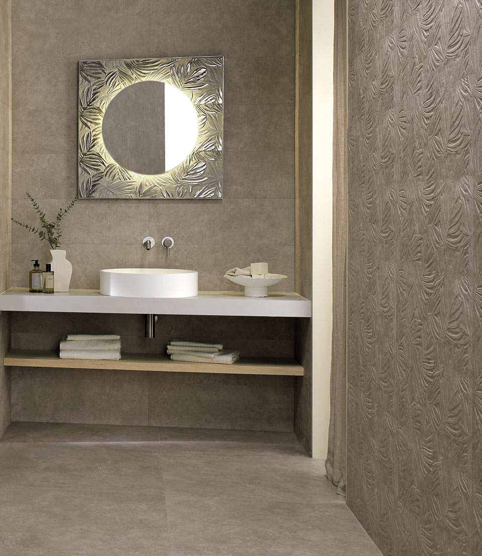 Bathroom with grey stone effect cladding and wall decorated in relief with a botanical motif, round mirror, white washbasin.