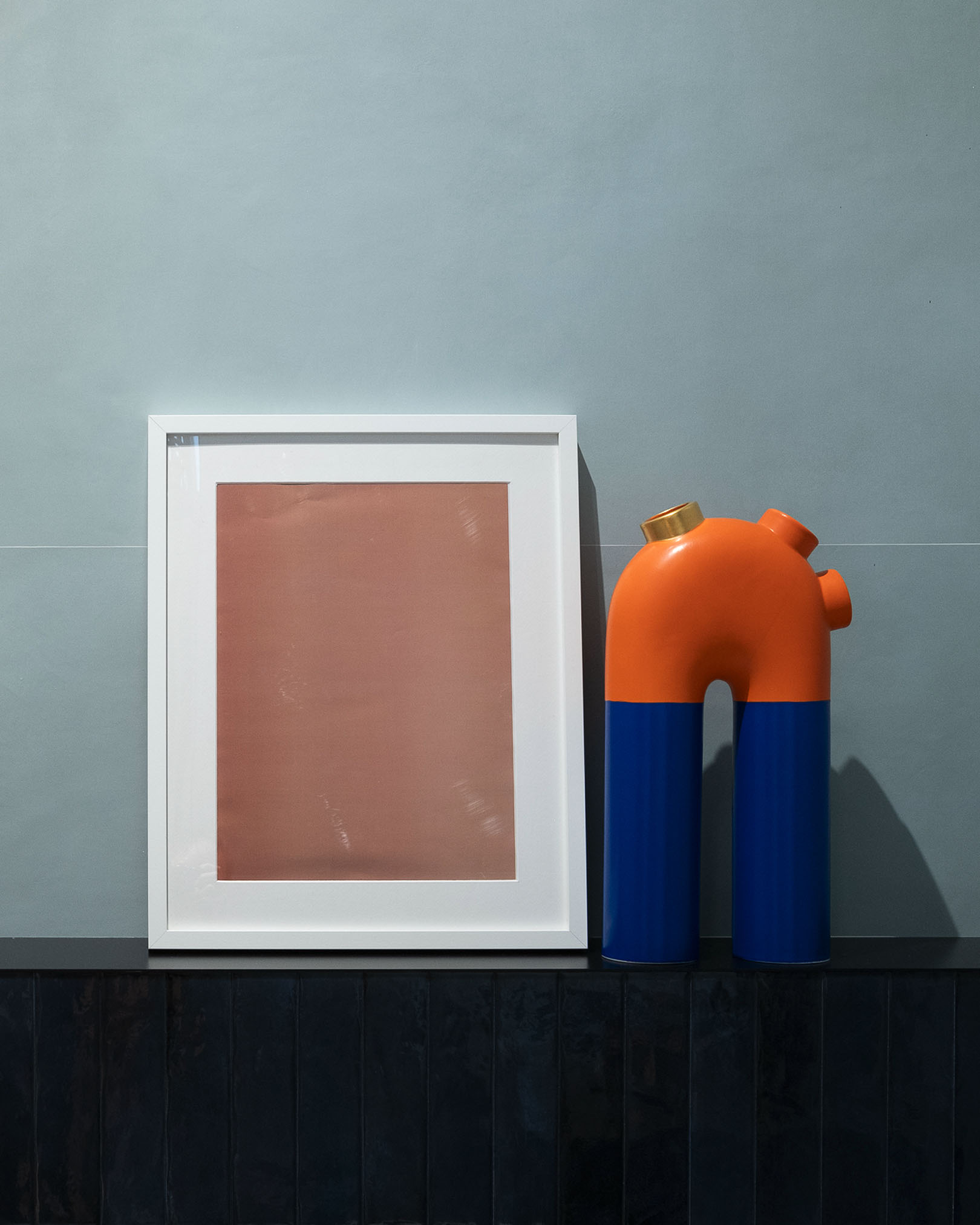 Grey wall with matt ceramic cladding, minimalist painting and orange-blue vase with a contemporary design on a dark base.
