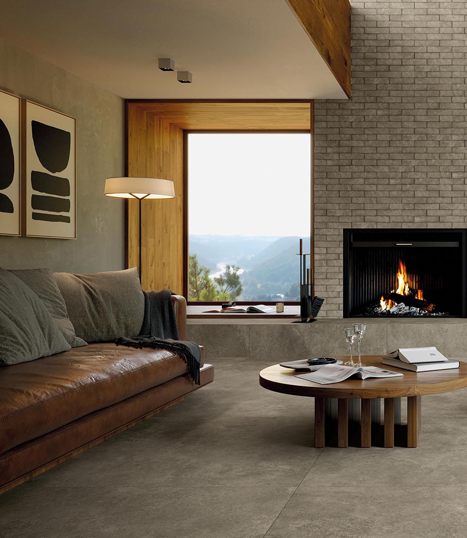 Modern living room with grey stone effect floor, grey brick walls, lighted fireplace, leather-coloured leather sofa, round wooden table.