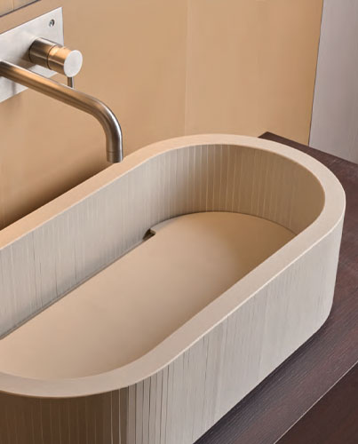 Oval washbasin in matt beige ceramic with vertical texture, dark wooden countertop and satin steel wall-mounted tap.