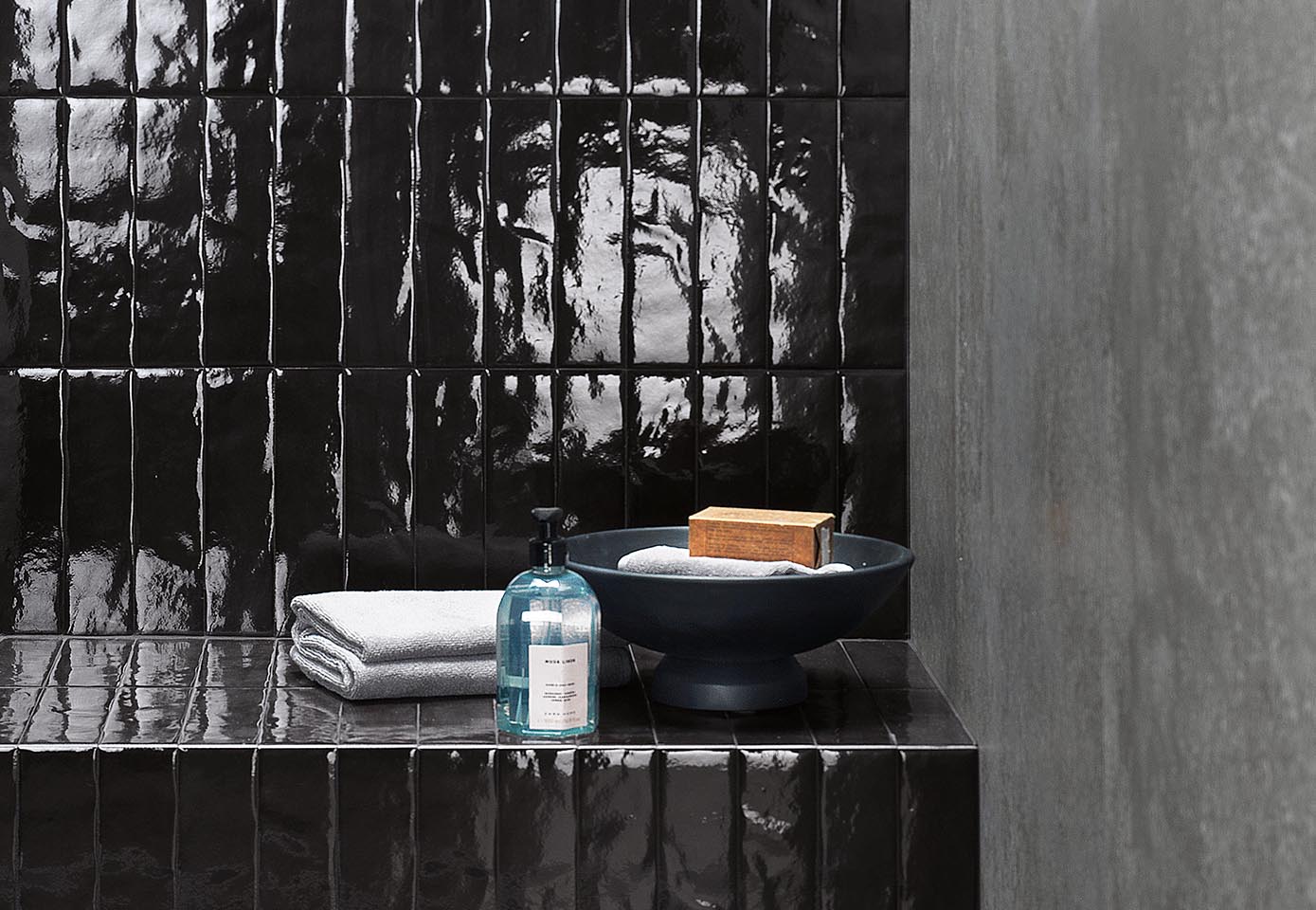 Detail of glossy black brick effect tiling, matt grey side wall, and various bathroom objects.