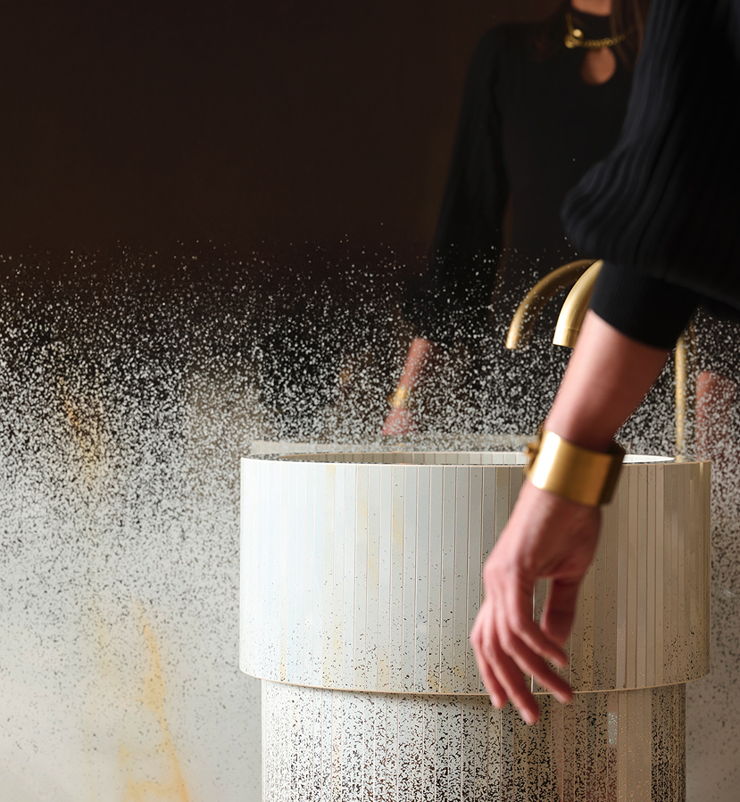 Detail of a washbasin decorated with a shaded mirror effect, a female arm with a gold bracelet, on a dark, enveloping background.