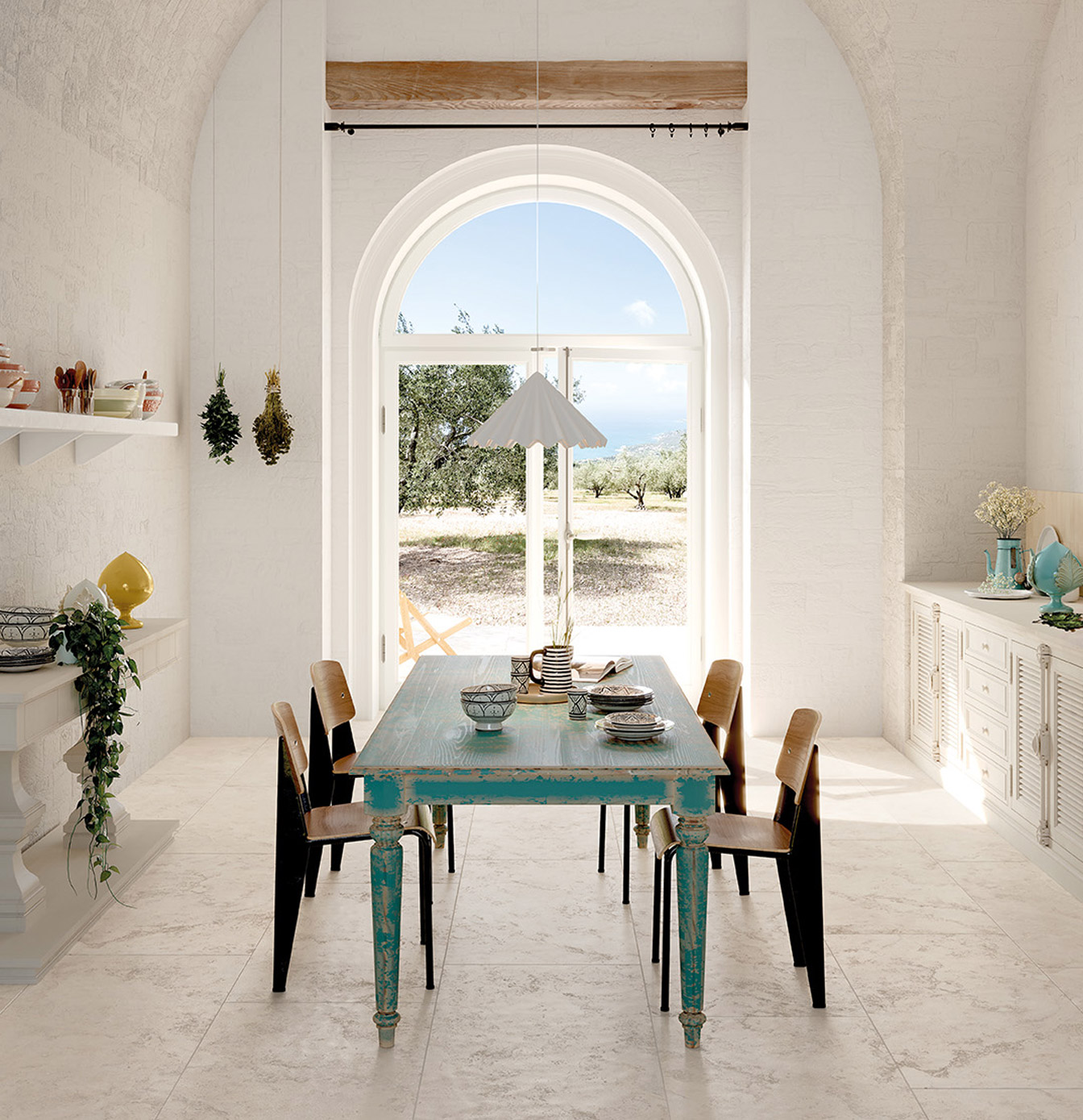 Bright Mediterranean-style kitchen with light stone effect floor, white textured walls and turquoise vintage table, black chairs with brown seats, various objects on shelves and cupboards.