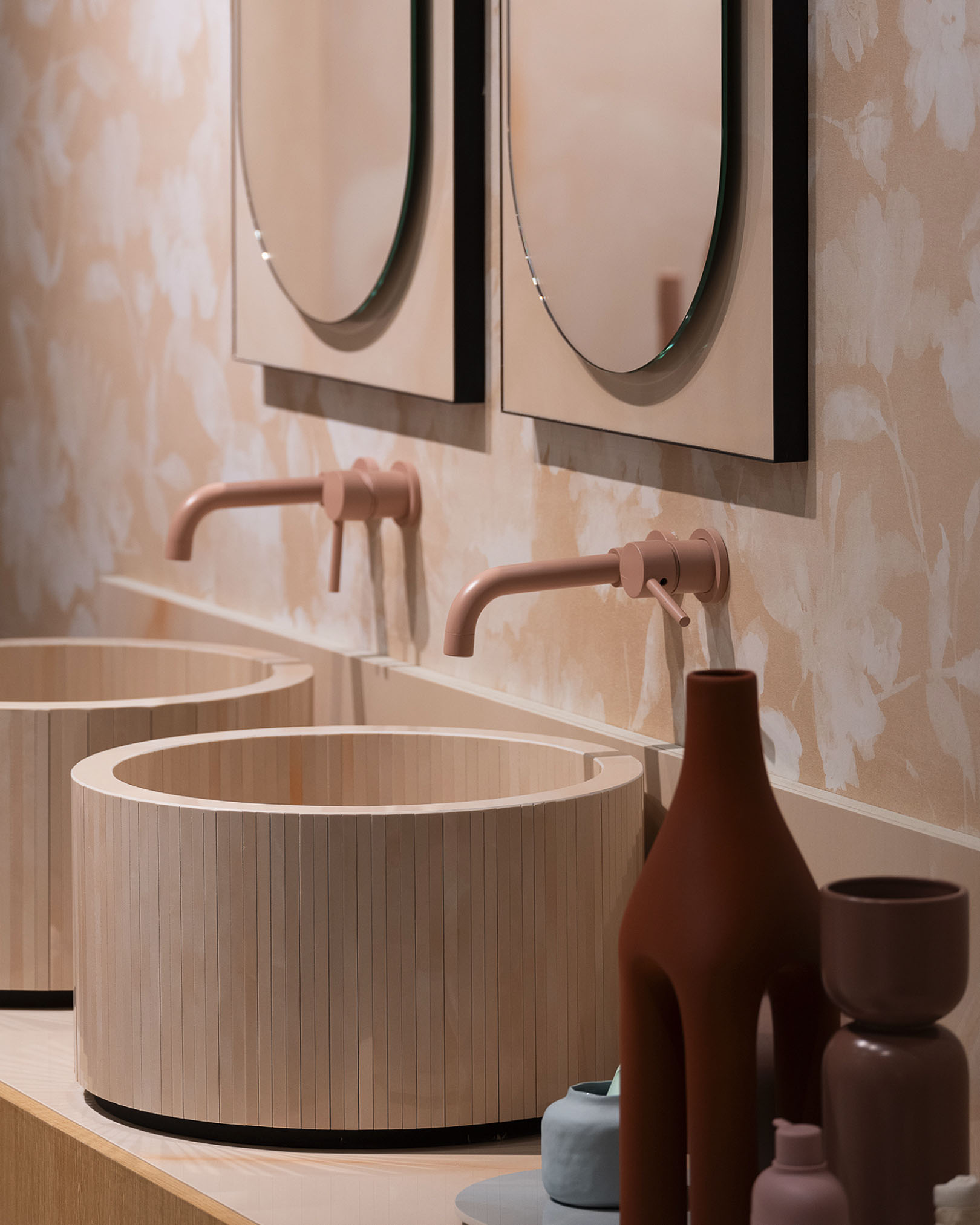 Bathroom with cylindrical light wood washbasin, matt pink taps and beige floral wall tiles, enriched with decorative vases.