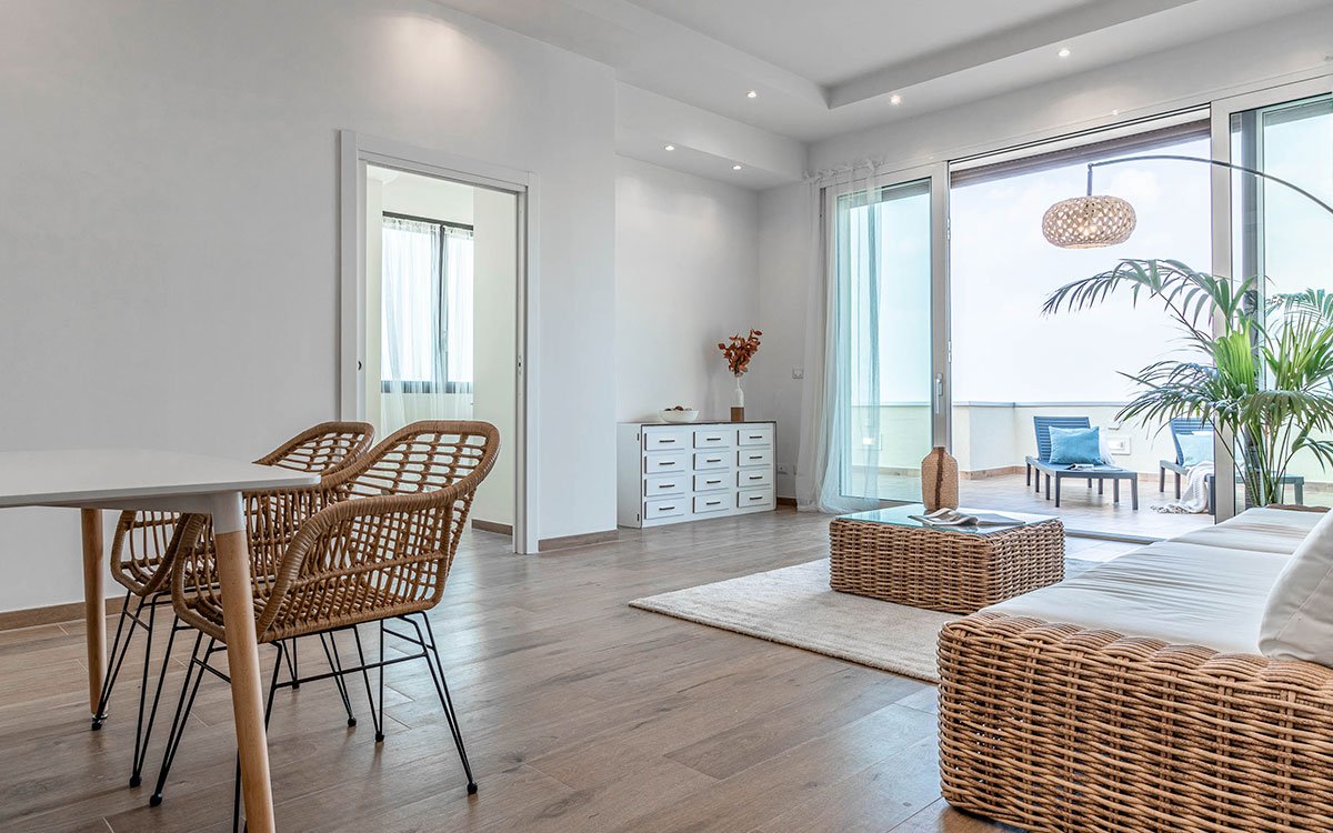 Soggiorno luminoso con pavimento effetto legno, arredi in rattan e vista terrazza arredata con chaise longue.