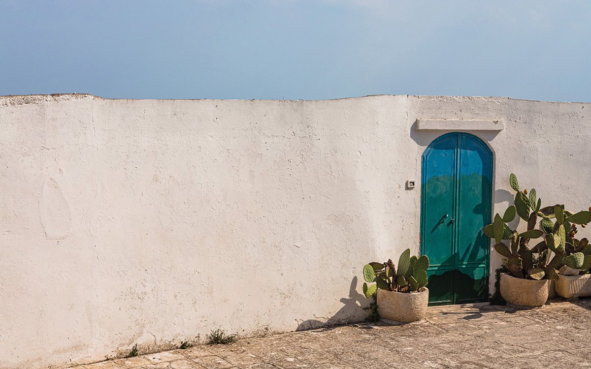 Muro bianco in calce con porta turchese e piante di fico d’India in vaso, ispirazione mediterranea autentica.