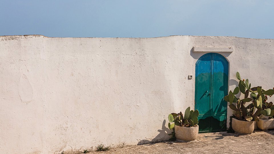 Muro bianco in calce con porta turchese e piante di fico d’India in vaso, ispirazione mediterranea autentica.