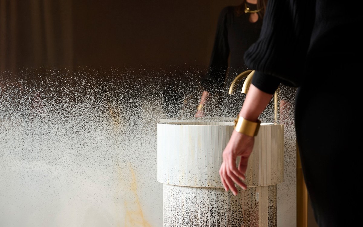 Cylindrical washbasin in light ceramic with gold tap, textured details and luminous reflections for a contemporary bathroom design.